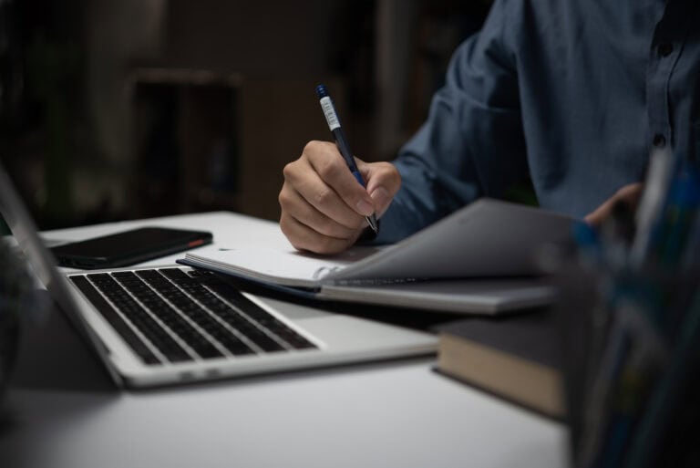 Billede af computer med en person som sidder og skriver i en notesbog